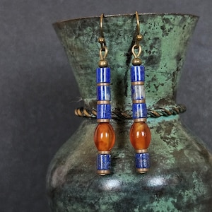 May include: A pair of dangle earrings featuring a vertical arrangement of cylindrical blue lapis lazuli beads and a single central oval orange carnelian bead, all connected by bronze-toned metal spacers and finished with bronze-toned ear hooks.