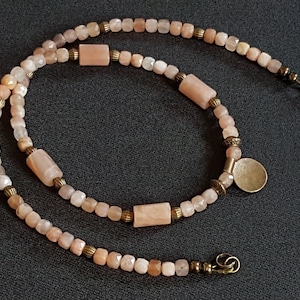 May include: A double-strand necklace made with peach-coloured sunstone beads and gold-toned metal accents. The necklace features a round pendant and a clasp.