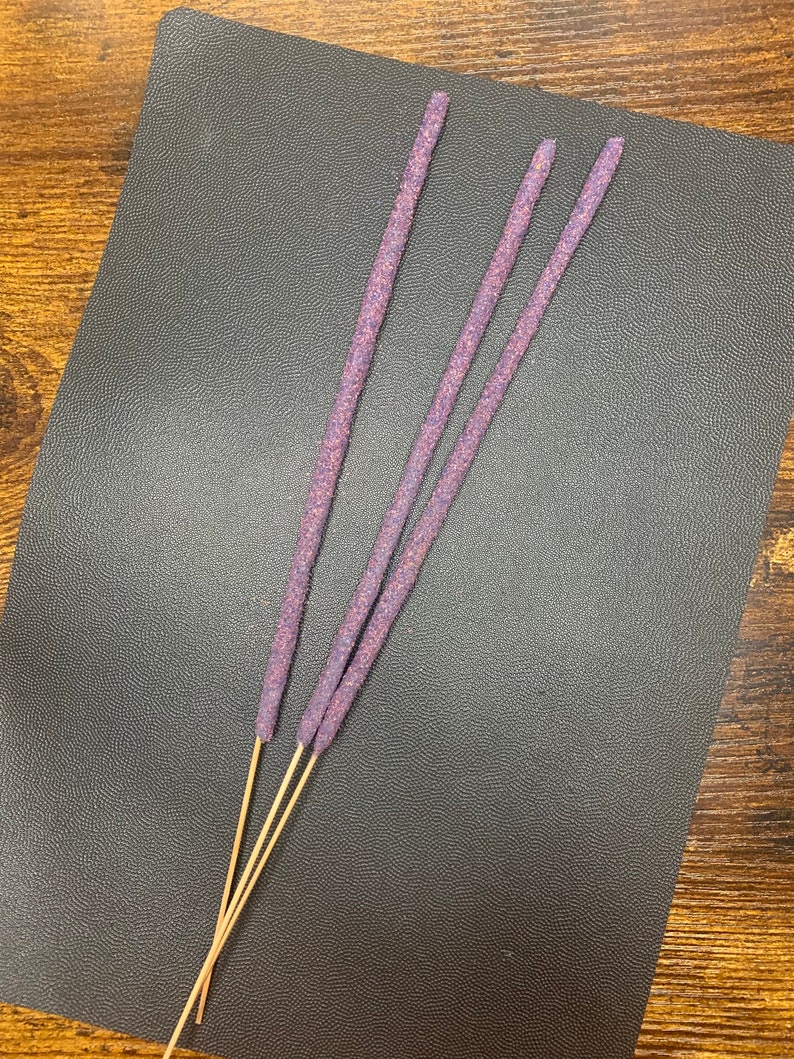 May include: Three purple incense sticks with wooden bases on a dark gray surface.