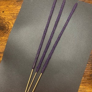 May include: Four purple incense sticks with a gold base on a dark gray surface.