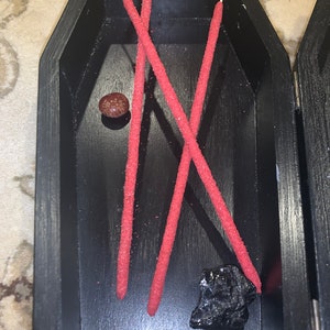 May include: Three red incense sticks and a small black stone in a black coffin-shaped box. The box is made of wood and has a hinged lid.