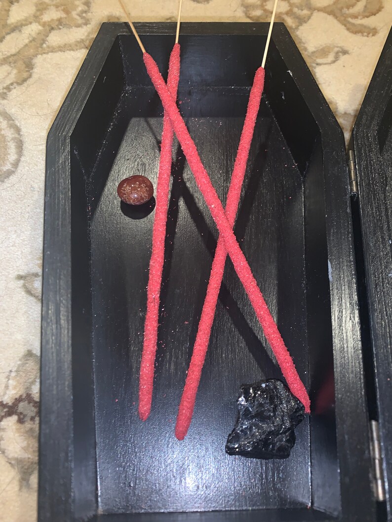May include: Three red incense sticks and a small black stone in a black coffin-shaped box. The box is made of wood and has a hinged lid.