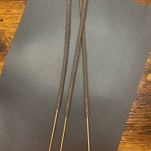 May include: Three brown incense sticks with a light brown wooden base on a dark gray textured surface.