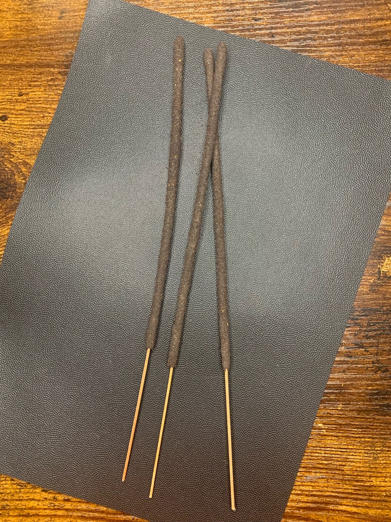 May include: Three brown incense sticks with a light brown wooden base on a dark gray textured surface.