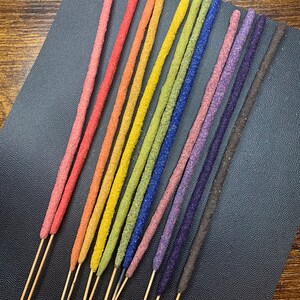 May include: A set of 12 incense sticks with a rainbow of colors, including red, orange, yellow, green, blue, purple, and brown. The sticks are arranged in a fan shape on a black surface.