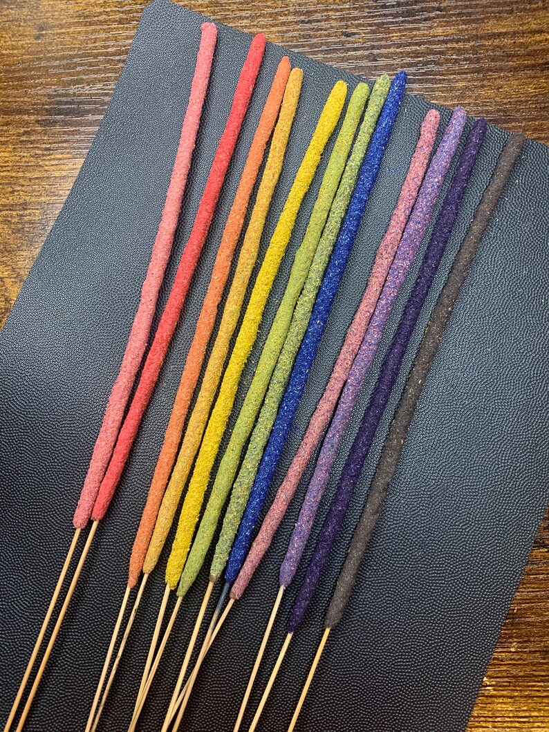 May include: A set of 12 incense sticks with a rainbow of colors, including red, orange, yellow, green, blue, purple, and brown. The sticks are arranged in a fan shape on a black surface.