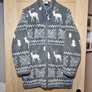 Op de afbeelding: Grijze fleecejas met een volledige ritssluiting, versierd met witte rendieren, sneeuwvlokken en boommotieven. De jas heeft een hoge kraag en een zachte textuur, ideaal voor koudere temperaturen.