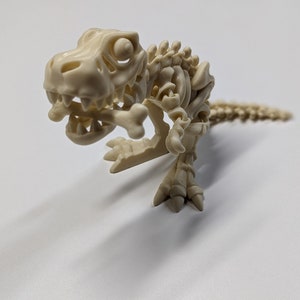 Puede incluir: Un juguete de esqueleto de dinosaurio de plástico blanco con un hueso en la boca. El dinosaurio está en posición de cuclillas con la cola enroscada alrededor de su cuerpo.