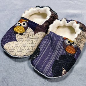 May include: A pair of baby booties with an owl design. The booties feature a purple, cream, and brown patterned fabric with a white interior. Each bootie has an owl applique with yellow and orange accents.