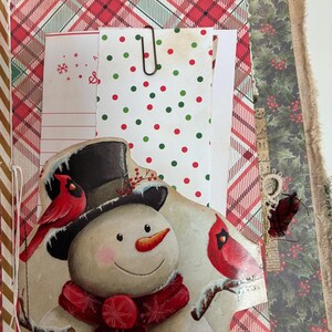 May include: A handmade journal page featuring a snowman illustration with a black top hat, a red scarf, and two red cardinals. The page includes a pocket with white paper containing red and green polka dots, held by a metal paperclip.