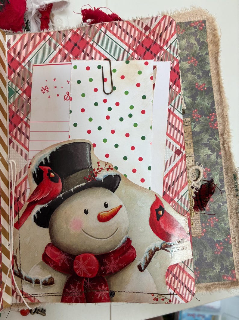May include: A handmade journal page featuring a snowman illustration with a black top hat, a red scarf, and two red cardinals. The page includes a pocket with white paper containing red and green polka dots, held by a metal paperclip.