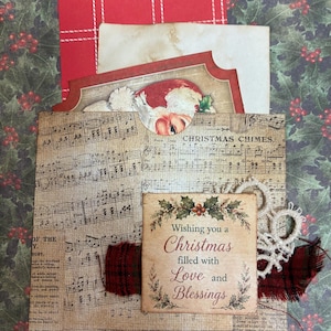 May include: A decorative paper pocket featuring vintage sheet music and the text Christmas Chimes. It holds a red plaid card, a cream-colored paper, and a Santa illustration. A square tag reads Wishing you a Christmas filled with Love and Blessings.