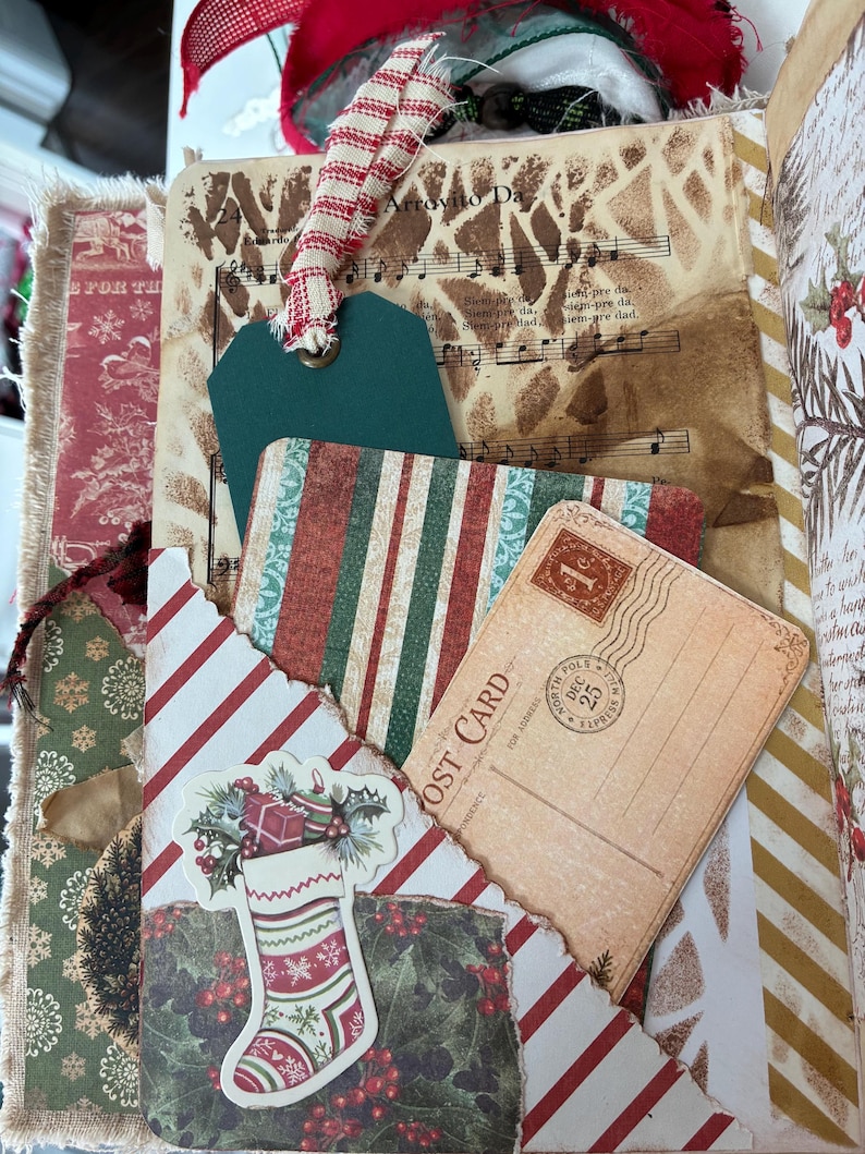 May include: A junk journal page featuring a vintage music sheet background, a dark green paper tag with a red and white striped ribbon, a striped card, and a postcard with a one-cent stamp. A die-cut Christmas stocking with holly is attached.