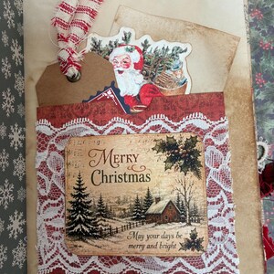 May include: A handmade journal page featuring a pocket with white lace trim. The pocket displays a winter scene with a cabin, pine trees, and the text Merry Christmas May your days be merry and bright. A Santa tag with red and white ribbon is tucked inside.