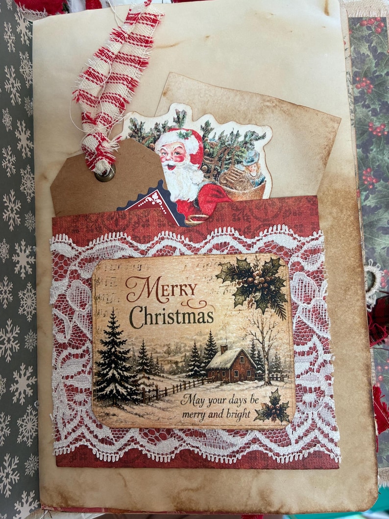May include: A handmade journal page featuring a pocket with white lace trim. The pocket displays a winter scene with a cabin, pine trees, and the text Merry Christmas May your days be merry and bright. A Santa tag with red and white ribbon is tucked inside.