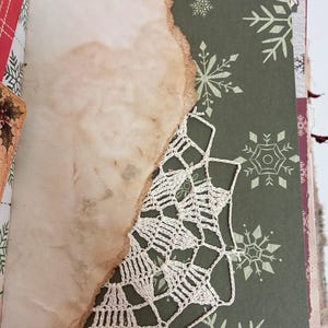 May include: A journal page featuring a dark green background with white snowflake patterns, partially covered by a piece of tea-stained paper with burnt, uneven edges. A white crocheted lace doily is layered between the paper and the page.