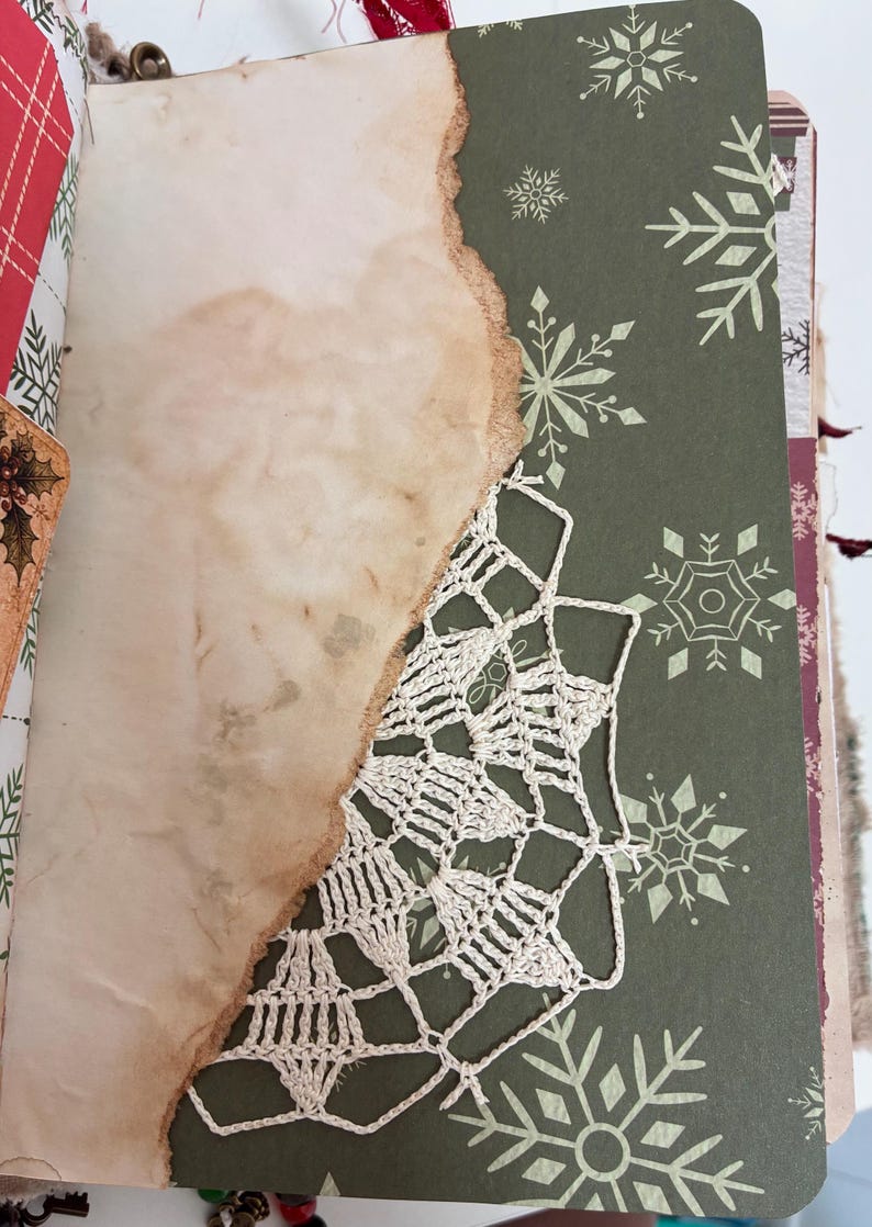 May include: A journal page featuring a dark green background with white snowflake patterns, partially covered by a piece of tea-stained paper with burnt, uneven edges. A white crocheted lace doily is layered between the paper and the page.