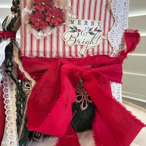 May include: A handmade journal with a red and white striped fabric cover featuring a Merry and Bright label, red poinsettia embellishments, white lace trim, and a large red fabric bow. The spine is decorated with hanging beads and ribbons.