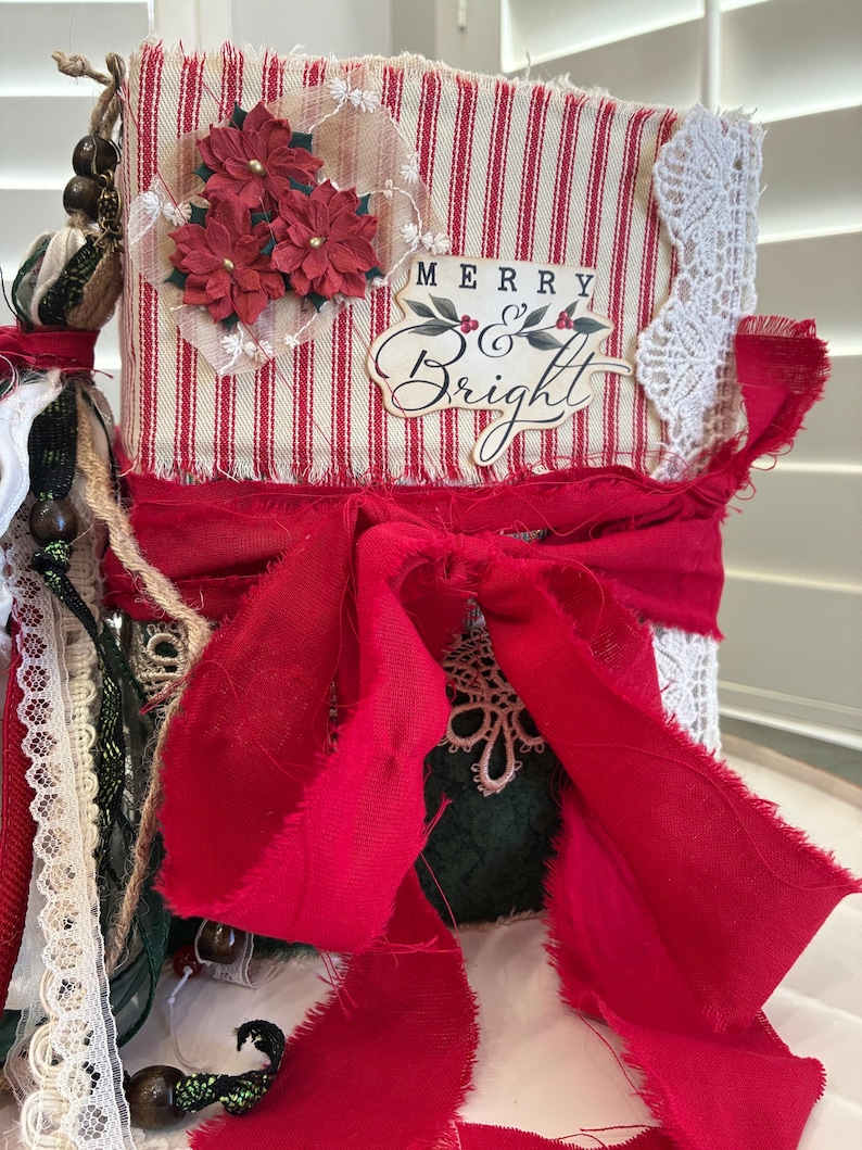 May include: A handmade journal with a red and white striped fabric cover featuring a Merry and Bright label, red poinsettia embellishments, white lace trim, and a large red fabric bow. The spine is decorated with hanging beads and ribbons.