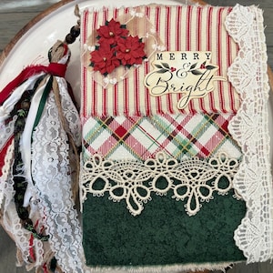 May include: A fabric-covered journal featuring a red and white striped top section with three red poinsettia flowers and the text Merry and Bright, a plaid middle section, and a dark green bottom section, accented with cream lace and a fabric tassel.
