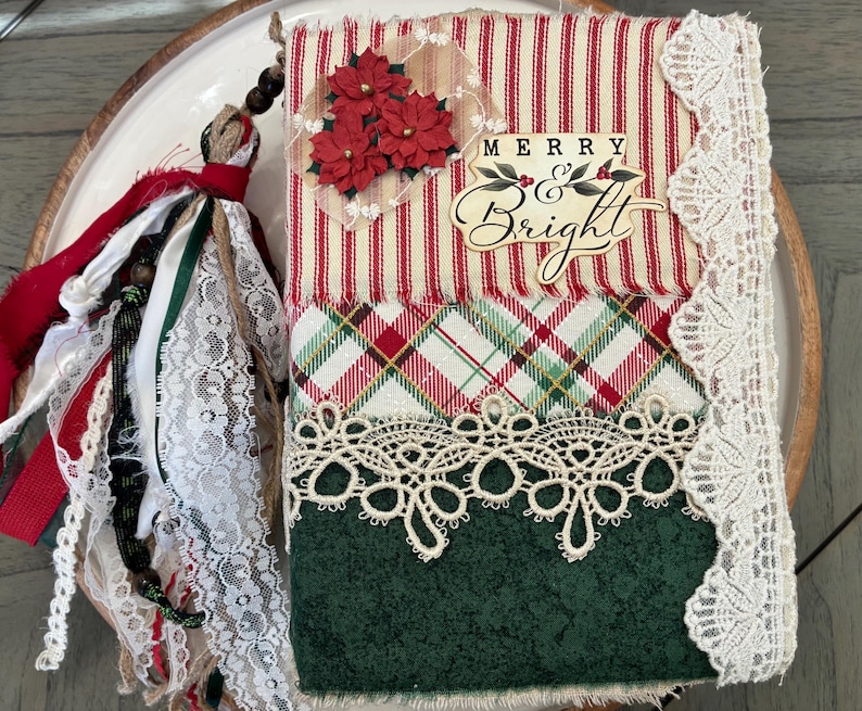 May include: A fabric-covered journal featuring a red and white striped top section with three red poinsettia flowers and the text Merry and Bright, a plaid middle section, and a dark green bottom section, accented with cream lace and a fabric tassel.