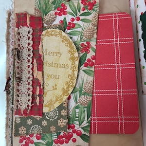 May include: A decorative paper tag featuring a pattern of green pine needles, brown pinecones, and red berries, topped with a red ribbon. It includes a circular cutout with the text Merry Christmas you, layered with red plaid paper, lace trim, and a safety pin.