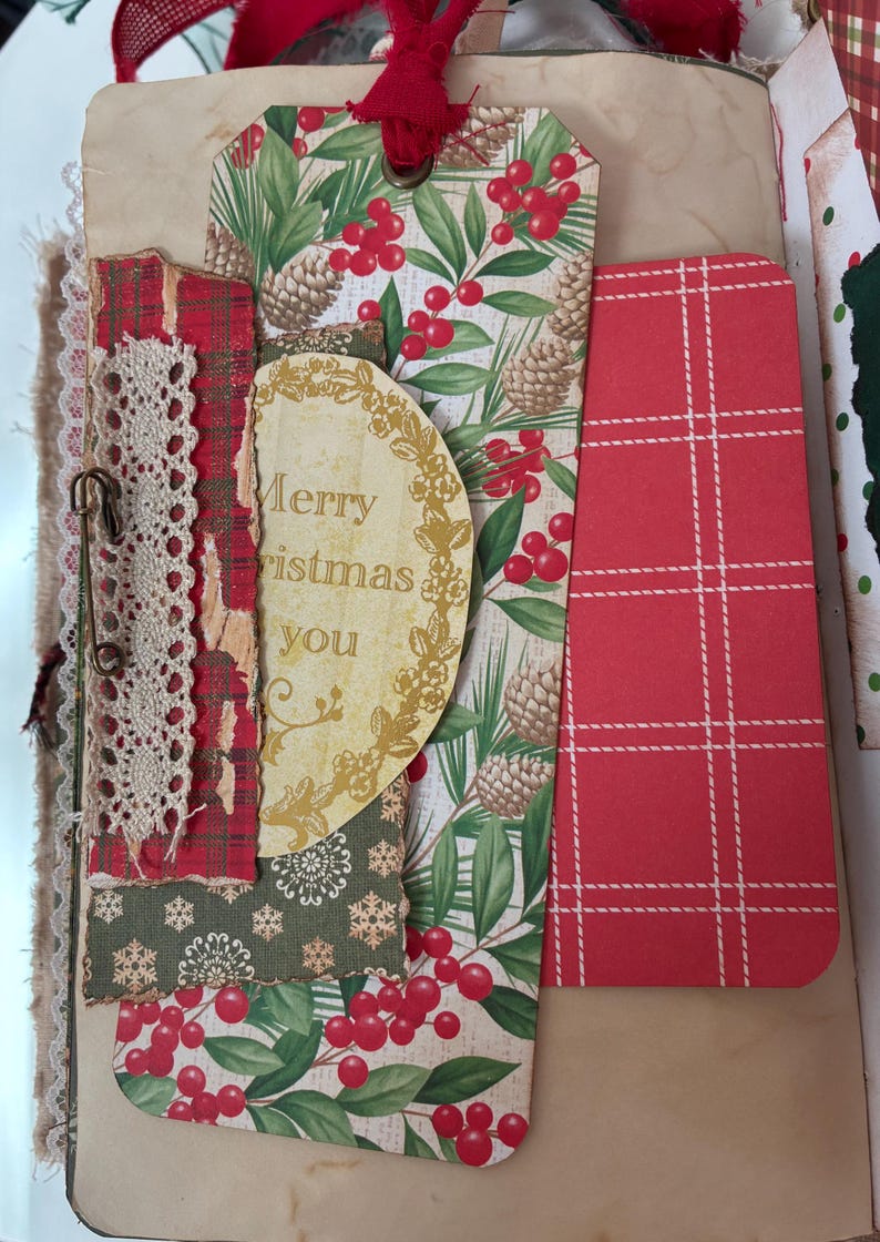 May include: A decorative paper tag featuring a pattern of green pine needles, brown pinecones, and red berries, topped with a red ribbon. It includes a circular cutout with the text Merry Christmas you, layered with red plaid paper, lace trim, and a safety pin.