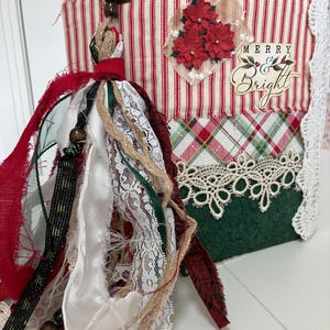 May include: A handmade journal featuring a red and white striped fabric cover with a plaid pattern, a green base, and white lace trim. The cover displays three red poinsettia flowers and the text Merry & Bright. A large fabric tassel hangs from the side.