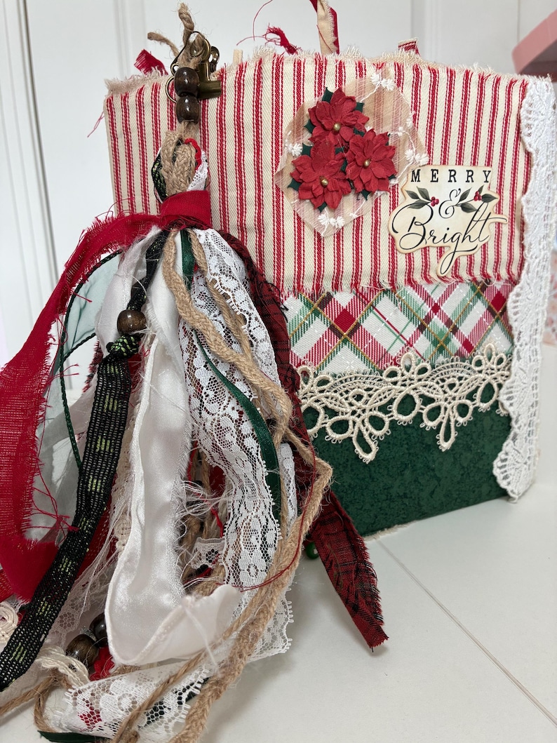 May include: A handmade journal featuring a red and white striped fabric cover with a plaid pattern, a green base, and white lace trim. The cover displays three red poinsettia flowers and the text Merry & Bright. A large fabric tassel hangs from the side.