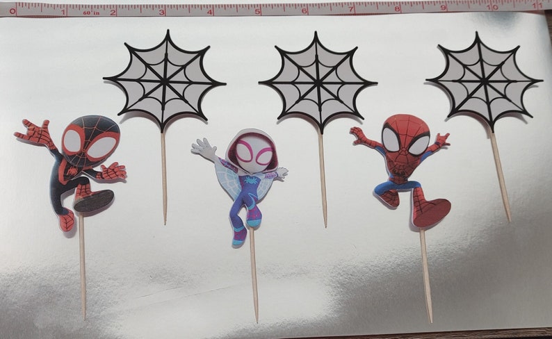 May include: Set of six cupcake toppers featuring Spider-Man characters. There are three black and white web designs, a red and black Spider-Man, a red and blue Spider-Man, and a white and purple Spider-Gwen.