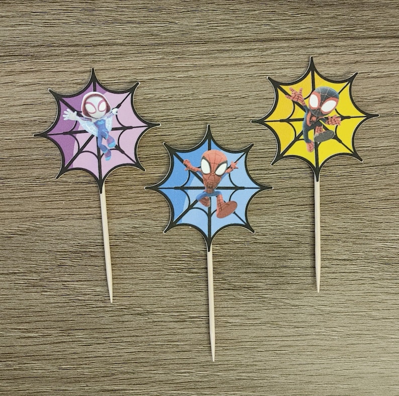 May include: Three Spider-Man themed cupcake toppers. Each topper features a different Spider-Man character in a web design. The toppers are on wooden sticks.