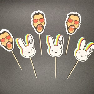 Puede incluir: Cinco adornos para cupcakes con un conejo arcoíris y un hombre con gafas de sol rojas en forma de corazón. Los adornos de conejo tienen el texto "Bad Bunny" en ellos. Los adornos de hombre tienen la lengua roja fuera.