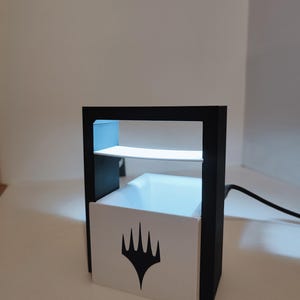 Puede incluir: Un expositor negro y blanco con una base blanca que presenta el logotipo de Wizards of the Coast. El expositor tiene un estante transparente y una luz en el interior.