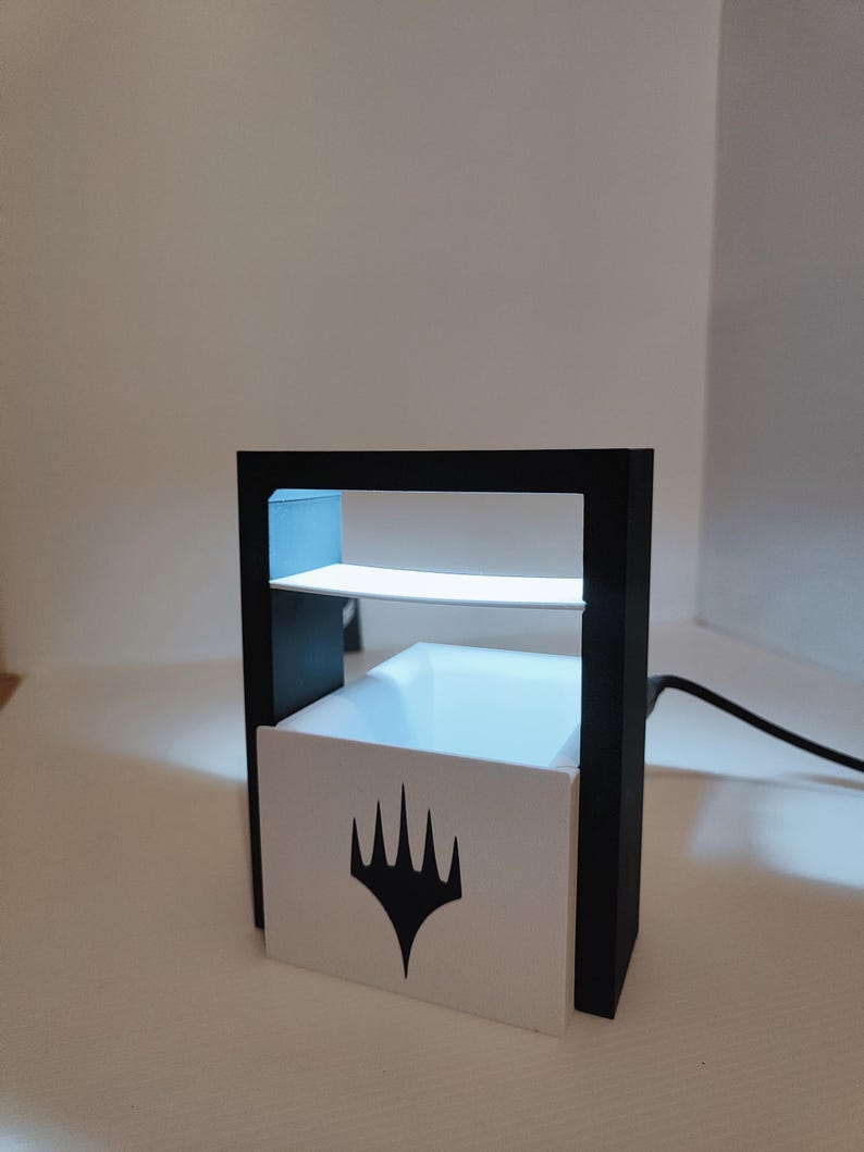 Puede incluir: Un expositor negro y blanco con una base blanca que presenta el logotipo de Wizards of the Coast. El expositor tiene un estante transparente y una luz en el interior.