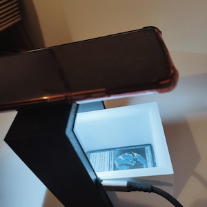 Puede incluir: Un soporte para tel&eacute;fono impreso en 3D con una luz blanca en su interior. Un tel&eacute;fono est&aacute; colocado en la parte superior del soporte y una tarjeta es visible dentro de la luz. El tel&eacute;fono est&aacute; conectado a un cable negro.