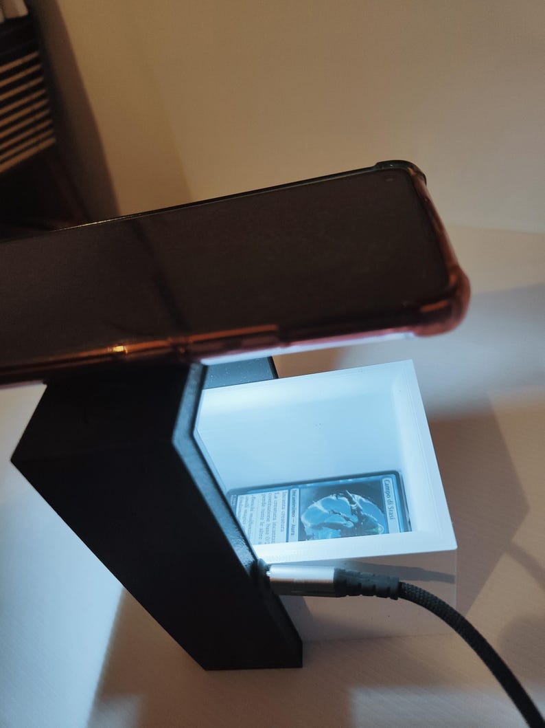 Puede incluir: Un soporte para tel&eacute;fono impreso en 3D con una luz blanca en su interior. Un tel&eacute;fono est&aacute; colocado en la parte superior del soporte y una tarjeta es visible dentro de la luz. El tel&eacute;fono est&aacute; conectado a un cable negro.