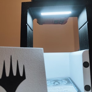 Puede incluir: Una caja blanca con un s&iacute;mbolo negro de Magic: The Gathering en la parte delantera. La caja est&aacute; abierta y tiene un interior blanco con una luz que brilla sobre ella. La caja est&aacute; sentada sobre una superficie negra.