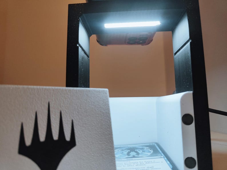 Puede incluir: Una caja blanca con un s&iacute;mbolo negro de Magic: The Gathering en la parte delantera. La caja est&aacute; abierta y tiene un interior blanco con una luz que brilla sobre ella. La caja est&aacute; sentada sobre una superficie negra.