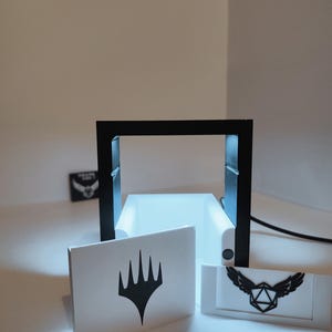Puede incluir: Un marco negro impreso en 3D con una base blanca y dos tarjetas blancas. La tarjeta de la izquierda tiene un s&iacute;mbolo negro de Magic: The Gathering. La tarjeta de la derecha tiene un s&iacute;mbolo negro de un dado alado.