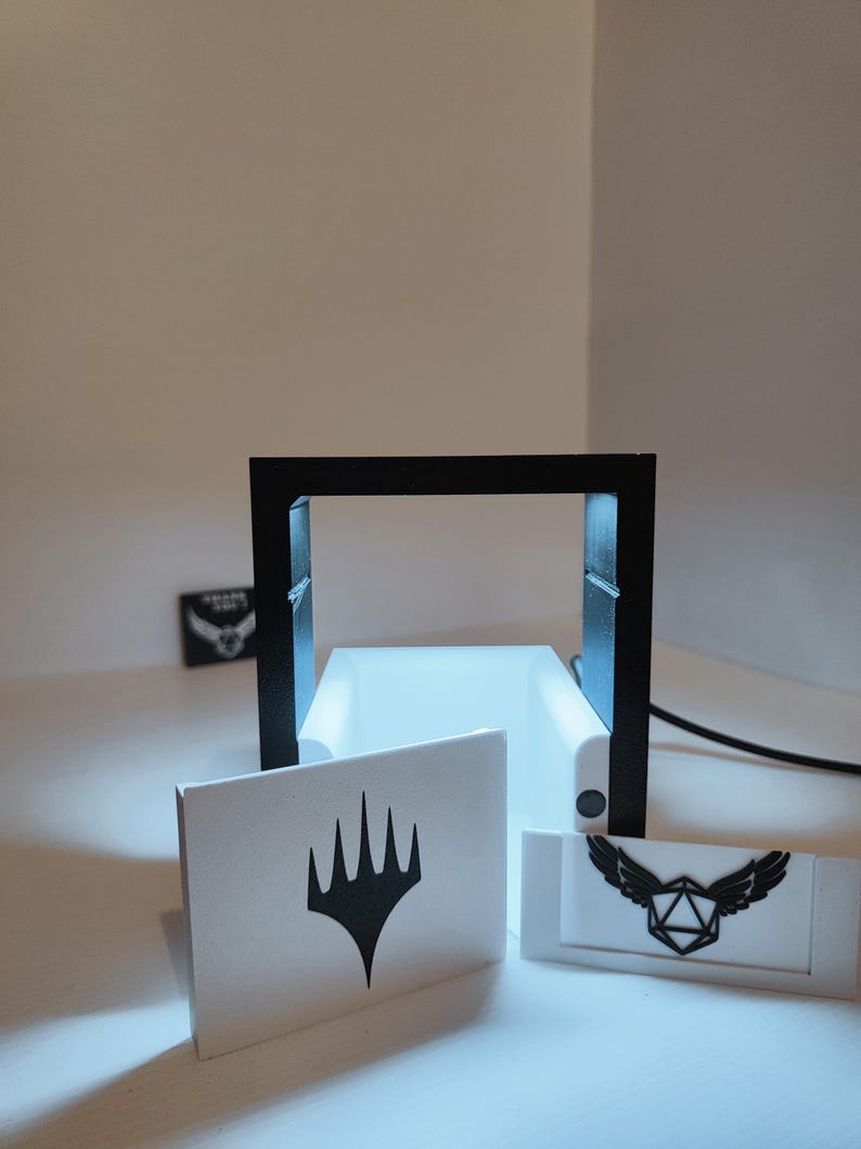 Puede incluir: Un marco negro impreso en 3D con una base blanca y dos tarjetas blancas. La tarjeta de la izquierda tiene un s&iacute;mbolo negro de Magic: The Gathering. La tarjeta de la derecha tiene un s&iacute;mbolo negro de un dado alado.