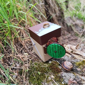 May include: A small wooden box with a green round door, resembling a hobbit hole, with a gold ring on top. The box is sitting on a brown log with green moss.
