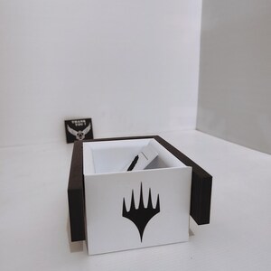 Puede incluir: Una caja blanca con bordes negros y un s&iacute;mbolo negro de Magic: The Gathering en la parte delantera. La caja est&aacute; vac&iacute;a.