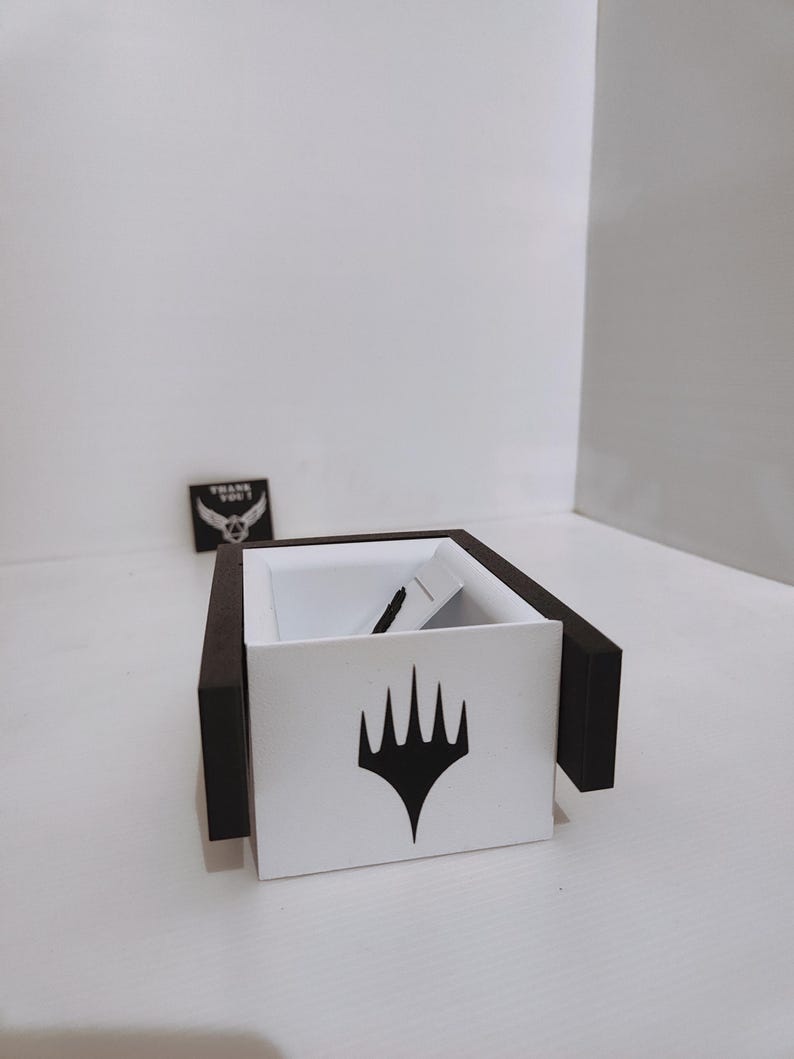 Puede incluir: Una caja blanca con bordes negros y un s&iacute;mbolo negro de Magic: The Gathering en la parte delantera. La caja est&aacute; vac&iacute;a.