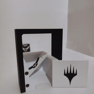 Puede incluir: Un barajador de cartas blanco y negro con una ventana transparente y un marco negro. El barajador tiene un portacartas blanco con un s&iacute;mbolo negro en &eacute;l. El barajador est&aacute; sentado sobre una superficie blanca.