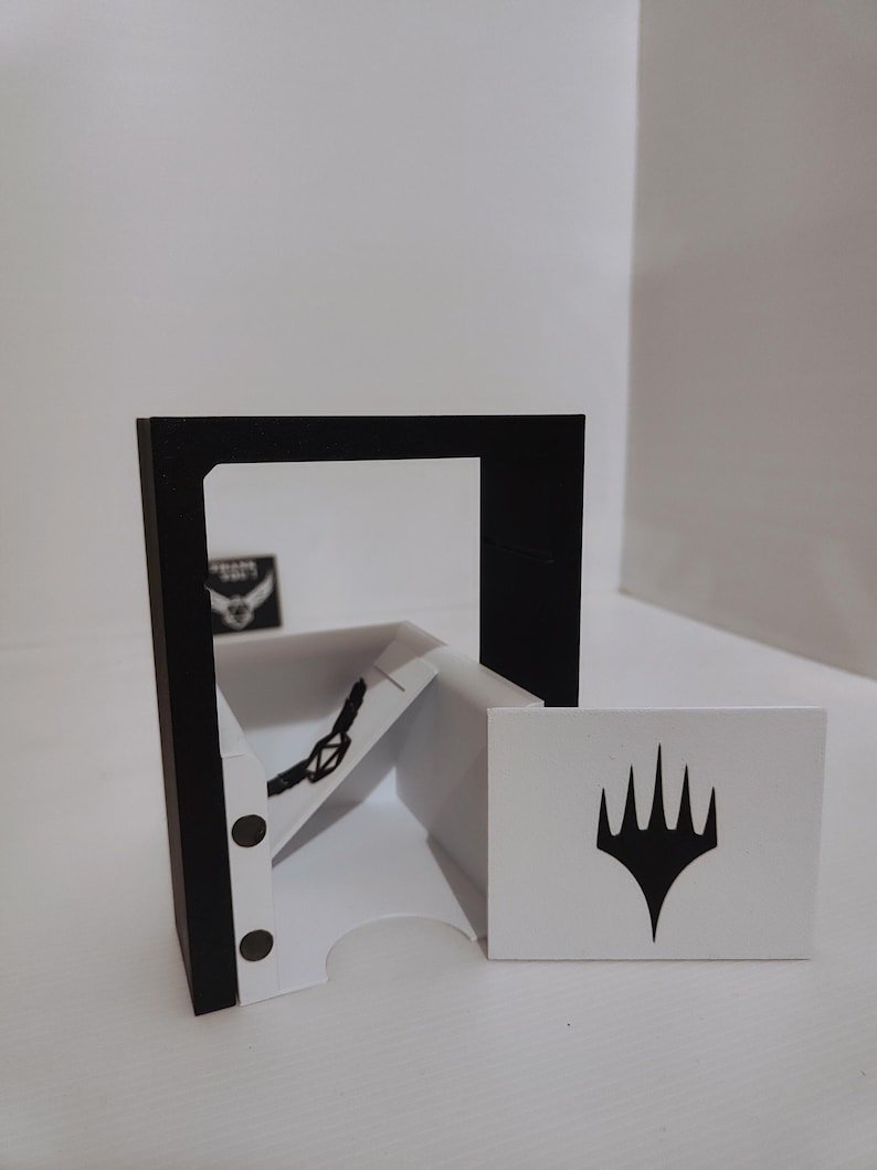 Puede incluir: Un barajador de cartas blanco y negro con una ventana transparente y un marco negro. El barajador tiene un portacartas blanco con un s&iacute;mbolo negro en &eacute;l. El barajador est&aacute; sentado sobre una superficie blanca.