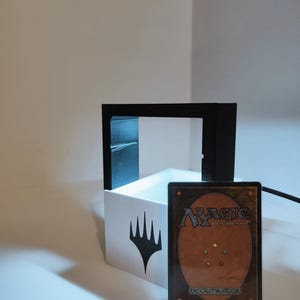 Puede incluir: Una caja blanca con un s&iacute;mbolo negro de Magic: The Gathering en la parte delantera. Un marco negro rodea la caja. Una carta de Magic: The Gathering con el texto "Deckmaster" est&aacute; delante de la caja.