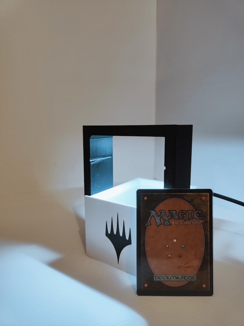 Puede incluir: Una caja blanca con un s&iacute;mbolo negro de Magic: The Gathering en la parte delantera. Un marco negro rodea la caja. Una carta de Magic: The Gathering con el texto "Deckmaster" est&aacute; delante de la caja.