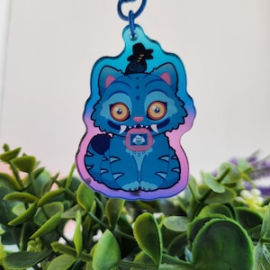 K&ouml;nnte beinhalten: Ein blauer, katzenf&ouml;rmiger Schl&uuml;sselanh&auml;nger mit Cartoon-Design. Die Katze hat gro&szlig;e orangefarbene Augen, einen kleinen Hut und ein kleines Bild auf der Brust. Der Schl&uuml;sselanh&auml;nger h&auml;ngt an einer blauen Schlaufe. Der Hintergrund ist eine Mischung aus Gr&uuml;n und Lila.