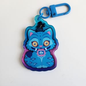 K&ouml;nnte beinhalten: Ein blauer, katzenf&ouml;rmiger Schl&uuml;sselanh&auml;nger mit einem blauen Verschluss. Die Katze hat gro&szlig;e gelbe Augen, einen kleinen rosa Mund und ein kleines Bild auf der Brust. Der Schl&uuml;sselanh&auml;nger hat einen blauen, lila und rosa Umriss.