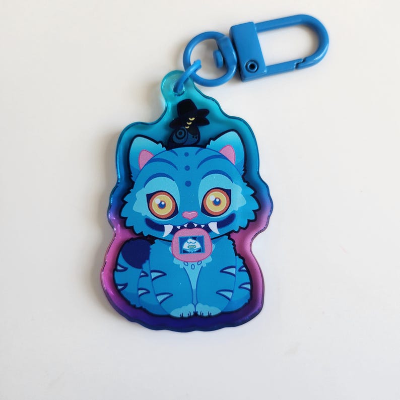 K&ouml;nnte beinhalten: Ein blauer, katzenf&ouml;rmiger Schl&uuml;sselanh&auml;nger mit einem blauen Verschluss. Die Katze hat gro&szlig;e gelbe Augen, einen kleinen rosa Mund und ein kleines Bild auf der Brust. Der Schl&uuml;sselanh&auml;nger hat einen blauen, lila und rosa Umriss.